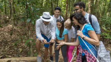 A group of smiling friends in casual clothes gather in a lush forest, looking at a smartphone held by one person. The scene conveys adventure and camaraderie.
